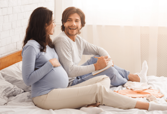 Futurs parents assis ensemble dans une chambre lumineuse, écrivant des idées de prénoms dans un carnet pour leur futur bébé.