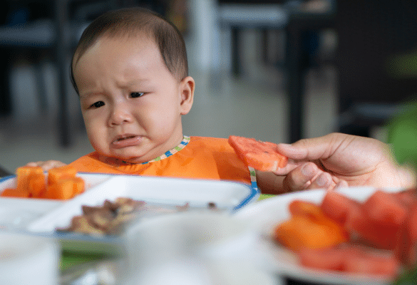Bébé assis dans sa chaise haute, regardant son assiette sans envie, pendant qu’un parent tente de le rassurer.