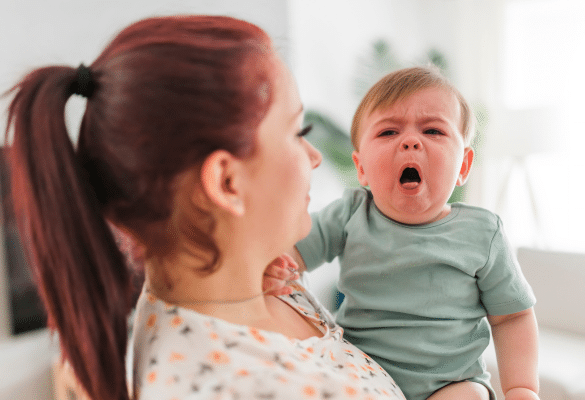 Bébé qui tousse et pleure dans les bras de sa mère, illustrant une toux pouvant provoquer des vomissements.