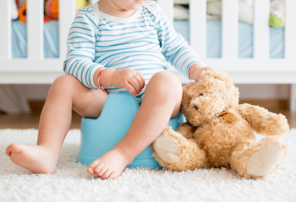 Bébé assis sur son pot avec un ours en peluche dans une chambre, symbolisant les débuts de l’apprentissage de la propreté.
