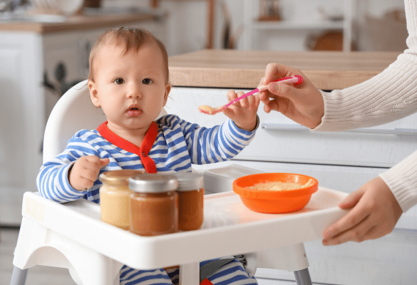 Bébé assis dans sa chaise haute, goûtant sa première purée de carotte avec un parent souriant à ses côtés