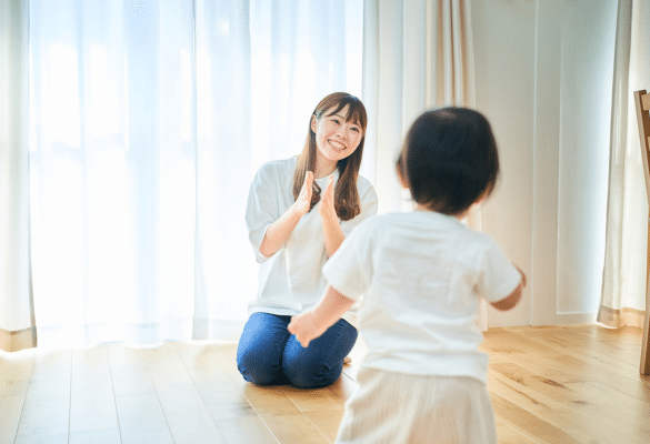 Parent à genoux face à un jeune enfant debout dans une pièce lumineuse ; le parent applaudit et invite l’enfant à imiter le geste dans un jeu de rythme.