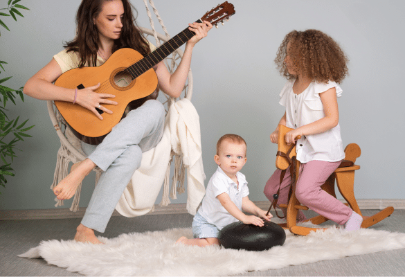 Parent assis sur un tapis jouant de la guitare pendant que deux jeunes enfants, assis et debout à proximité, écoutent et participent dans une ambiance douce et détendue.