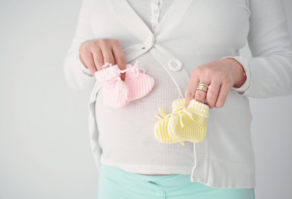 Femme enceinte tenant une paire de chaussons roses et bleus sur son ventre rond, symbolisant l’attente de jumeaux.