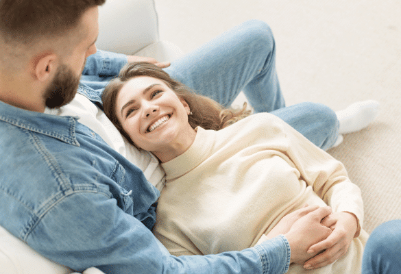 Femme enceinte souriante allongée contre son partenaire sur un canapé, dans un moment de complicité