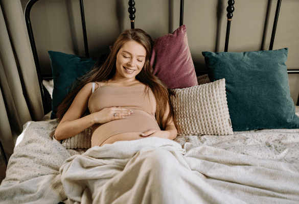 Femme enceinte souriante assise dans son lit, les mains posées sur son ventre arrondi